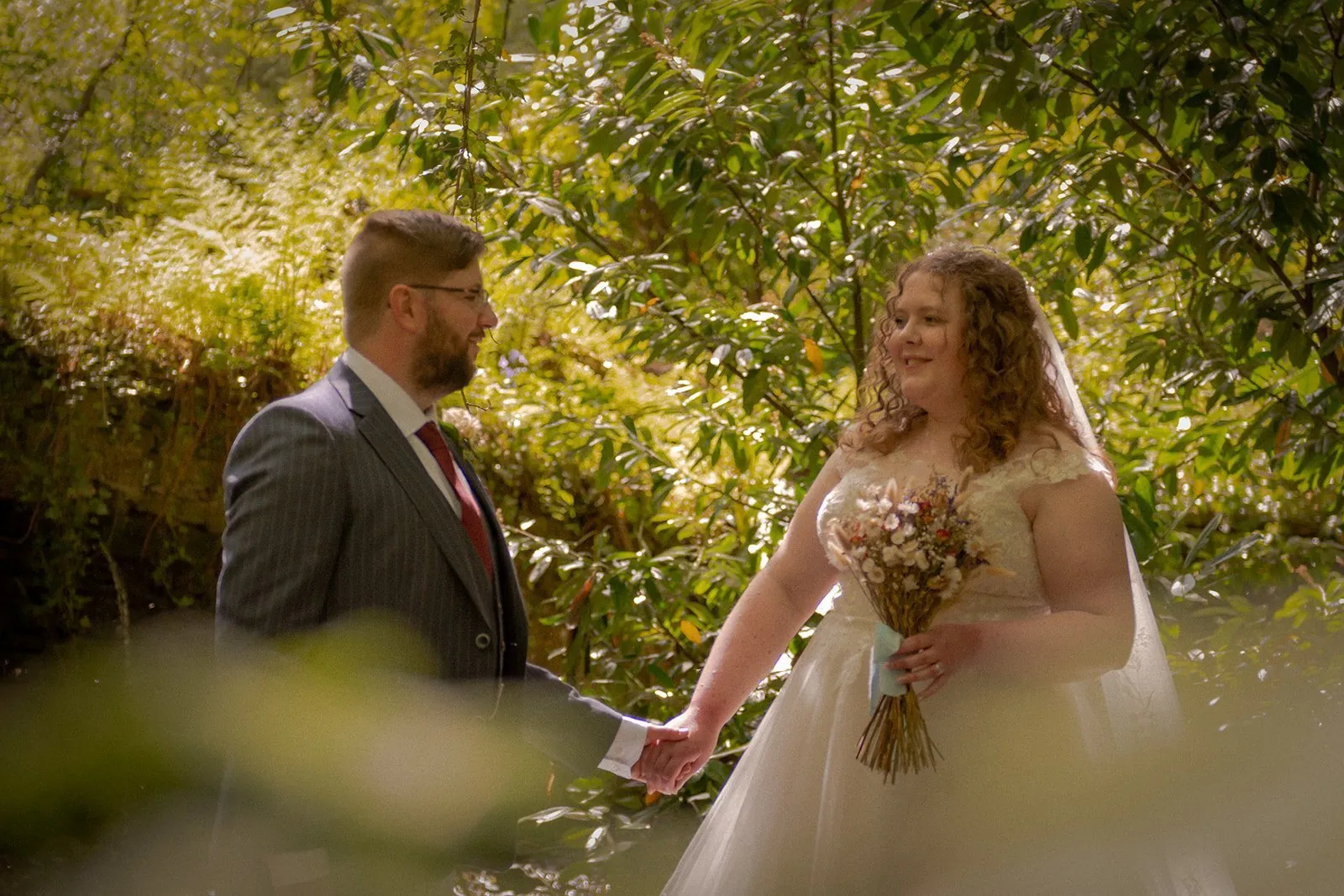 Bride and groom holding hands in dappled woodland light, the bride carrying a loose garden-flower bouquet